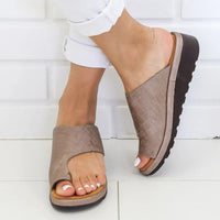 Sandale Nue Pied Femme Design Style Romain portée par une femme avec un pantalon blanc devant un mur en brique blanche