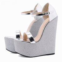 Sandale argentée et pailletée style plateforme. La chaussure présente une semelle plateforme ultra-épaisse en paillettes grises scintillantes avec finition noire à la base, un talon compensé assorti très volumineux, des lanières blanches métallisées brillantes, une bride de cheville avec boucle carrée argentée réglable, design très festif et spectaculaire. Présentée sur fond blanc.