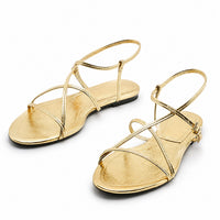 Sandale dorée avec lanières fines et élégantes. La chaussure présente un design métallisé doré brillant, des lanières croisées fines en T qui encadrent le pied, une bride arrière ajustable avec boucle dorée, et une semelle plate fine. Présentée sur fond blanc.