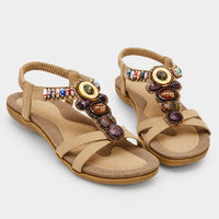 Sandale femme beige de style bohème avec lanières. La chaussure présente une semelle plate confortable en cuir beige-taupe, des lanières croisées en cuir beige, une décoration très ornée sur la bride de pied avec médaillons dorés en cabochons, perles multicolores, pierre d'améthyste et œil de tigre, perles blanches et colorées, finitions dorées sophistiquées. Une bride arrière enveloppante très confortable. Design très ethnique et bohème. Présentée sur fond blanc.