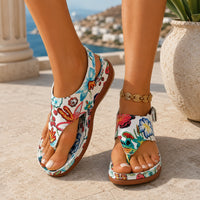 Sandale femme confortable style tong avec motif floral. La chaussure présente une semelle plate confortable en caoutchouc marron-caramel, une bande tong avec motifs floraux très colorés sur fond blanc-crème aux teintes bleu, rouge, orange, jaune et vert, design très décontracté et printanier. Portée sur une terrasse méditerranéenne ensoleillée avec vue sur la mer turquoise.