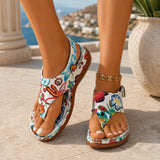 Sandale femme confortable style tong avec motif floral. La chaussure présente une semelle plate confortable en caoutchouc marron-caramel, une bande tong avec motifs floraux très colorés sur fond blanc-crème aux teintes bleu, rouge, orange, jaune et vert, design très décontracté et printanier. Portée sur une terrasse méditerranéenne ensoleillée avec vue sur la mer turquoise.
