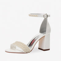 Sandale femme mariage blanche avec perles sur les lanières. La chaussure présente un talon carré blanc de hauteur moyenne, un bout ouvert, une lanière supérieure densément ornée de perles blanches nacrées, une bride de cheville avec boucle argentée réglable, une semelle doublée métallisée argent. Design très luxueux et délicat pour mariage, présenté sur fond blanc.