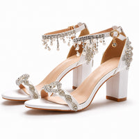 Sandale femme mariage blanche avec strass et perles suspendues. La chaussure présente un talon carré blanc de hauteur moyenne, des bandes strass brillants ornant le devant du pied en motifs géométriques, une bride de cheville blanche décorée de strass, et une frange de perles blanches suspendues qui retombent élégamment. Design très luxueux et sophistiqué, présenté sur fond blanc.