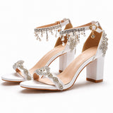 Sandale femme mariage blanche avec strass et perles suspendues. La chaussure présente un talon carré blanc de hauteur moyenne, des bandes strass brillants ornant le devant du pied en motifs géométriques, une bride de cheville blanche décorée de strass, et une frange de perles blanches suspendues qui retombent élégamment. Design très luxueux et sophistiqué, présenté sur fond blanc.