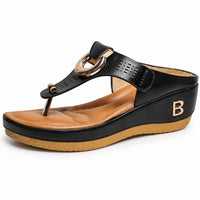 Sandale femme moderne avec semelle et lanières épaisses noir-beige. La chaussure présente une semelle plateforme très épaisse bicolore noir et taupe-beige avec profil texturé, une bande tong noire ornée de découpes perforées ajourées, un détail ornemental doré sur la bande tong, une fermeture en T avec boucle dorée, doublure marron-caramel contrastante, initial 