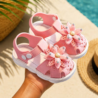 Sandale fille antidérapante et respirante rose. La chaussure présente des fleurs roses à paillettes avec perles dorées et strass sur le dessus, une semelle blanche aérée avec trous de drainage, et une bride arrière ajustable. Présentée sur fond de plage ensoleillée près d'une piscine.