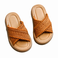 Sandale fille style tong avec lanières tressées et croisées caramel. La chaussure présente une semelle plate confortable en caoutchouc beige clair avec profil arrondi, une bande tong en tissu tressé caramel-abricot, des lanières croisées ornées de lanières tressées multicolores avec petits détails de pompons/boules tout autour des bandes pour un effet très ludique et mignon, doublure intérieure beige très confortable, design très mignon adapté aux enfants. Présentées sur fond blanc.