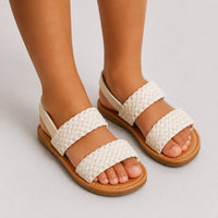 Sandale fille avec lanières tressées beige-crème. La chaussure présente une semelle plate confortable en cuir marron-caramel clair, des lanières très larges tressées/tissées en coton beige-crème très douces avec finitions arrondies épaisses très confortables, un design très minimaliste et naturel adapté aux enfants, bride arrière simple en cuir beige-crème, style très bohème et décontracté. Très confortable et chic pour enfant. Portée sans chaussettes sur fond neutre.