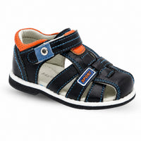 Sandale garçon plate en simili-cuir noir. La chaussure présente une semelle plate blanche contrastante, un corps en simili-cuir noir avec accents orange et détails bleu ciel, une fermeture velcro/scratch réglable avec panneau denim bleu ciel et œillet métallique, découpes ajourées latérales pour l'aération, doublure intérieure grise très confortable, coutures décoratives bleu ciel tout autour, badge 