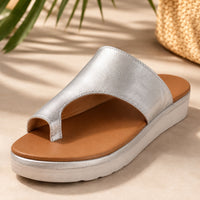 Sandale nu-pied femme design style romain argent-caramel. La chaussure présente une semelle plateforme plate blanche-crème très confortable avec profil arrondi, une bande tong métallisée argent brillant très minimaliste, une partie supérieure du pied en cuir caramel-marron très structuré, un design très épuré inspiré du style romain ancien avec découpes géométriques caractéristiques, doublure intérieure caramel contrastante, style très chic et intemporel. Présentée sur un sol beige ensoleillé avec plantes d