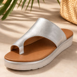 Sandale nu-pied femme design style romain argent-caramel. La chaussure présente une semelle plateforme plate blanche-crème très confortable avec profil arrondi, une bande tong métallisée argent brillant très minimaliste, une partie supérieure du pied en cuir caramel-marron très structuré, un design très épuré inspiré du style romain ancien avec découpes géométriques caractéristiques, doublure intérieure caramel contrastante, style très chic et intemporel. Présentée sur un sol beige ensoleillé avec plantes d