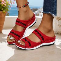 Sandale orthopédique femme rouge avec motif sur les lanières. La chaussure présente une semelle compensée épaisse avec base beige et bande blanche, des bandes croisées rouges en tissu texturé ornées de motifs géométriques noirs en zigzag, design confortable et supportif. Portée sur une terrasse méditerranéenne ensoleillée avec vue sur la mer.