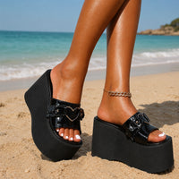 Sandale plateforme femme avec un cœur argenté noire. La chaussure présente une semelle plateforme ultra-épaisse en caoutchouc noir très volumineux, une bande de pied noire avec un détail ornamental en forme de cœur argenté laqué brillant au centre, design très tendance et ludique. Portée à la plage avec bracelet chaîne dorée, sur un sable blanc avec vue sur la mer turquoise en arrière-plan.