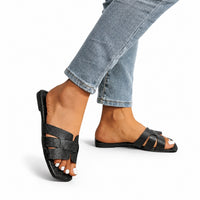 Sandale style tong noire et design. La chaussure présente une semelle plate en caoutchouc noir, une bande tong noire avec texture mate, des lanières noires croisées et minimalistes formant un design épuré très tendance, style très simple et contemporain. Portée avec un jean bleu clair, design confortable et casual.