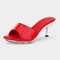 Sandale talon femme 5-7 cm rouge avec semelle transparente. La chaussure présente un talon transparent en acrylique cristal d'environ 5-7 cm de hauteur, une semelle plate transparente en acrylique cristal, une bande de pied/bout ouvert en cuir laqué rouge brillant, design très minimaliste et moderne avec effet cristal transparent très élégant. Portée dans un intérieur moderne avec miroir et éclairage LED en arrière-plan.