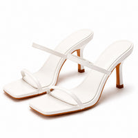 Sandale talon femme 7-10 cm blanche avec lanières fines. La chaussure présente un talon élevé fin blanc avec finition bois marron contrastant, une semelle plate carrée blanche avec bordure bois, des lanières minimalistes blanches croisées sur le dessus du pied, design très épuré et moderne. Présentée sur fond blanc.