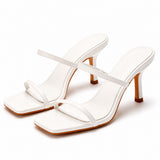 Sandale talon femme 7-10 cm blanche avec lanières fines. La chaussure présente un talon élevé fin blanc avec finition bois marron contrastant, une semelle plate carrée blanche avec bordure bois, des lanières minimalistes blanches croisées sur le dessus du pied, design très épuré et moderne. Présentée sur fond blanc.