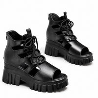 Sandale talon plateforme semi-ouverte avec lacets noire. La chaussure présente une semelle plateforme ultra-épaisse en caoutchouc noir texturé avec motifs de rainures profilées très volumineux, un talon compensé très épais noir assorti, un design semi-ouvert avec découpes ajourées latérales, des lacets noirs pour l'ajustement, plusieurs bandes noires de maintien, une fermeture arrière réglable, doublure intérieure noire, design très moderne avant-garde et tendance. Présentées sur fond blanc.