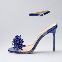 Sandale Bleu Marine Ouverte avec Lanière à Boucle avec finition brillante, ornement bleu et doré sur l’avant, bride fine réglable à la cheville, bout ouvert et talon aiguille élégant.