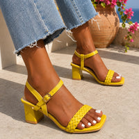Sandale à talon femme confortable et tendance jaune vif. La chaussure présente un talon carré jaune de hauteur moyenne, une semelle plate jaune, une bande tressée/entrelacée jaune ornant le devant du pied, une bride de cheville réglable avec boucle dorée. Design très coloré et moderne. Portée avec un jean bleu délavé sur une terrasse méditerranéenne ensoleillée avec fleurs fuchsia en arrière-plan.