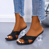 Sandale à talon femme avec lanières croisées et tressées noires. La chaussure présente un talon très original en acrylique transparent et géométrique, une semelle plate noire, des lanières tressées/entrecroisées en noir formant un motif entrelacé sur le dessus du pied, bout ouvert peep toe, design très moderne et tendance. Portée avec un jean bleu clair déchiré aux genoux.