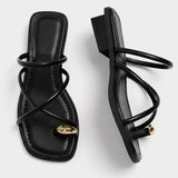 Sandales Plates Avec Brides Entrelacées Minimalistes noires avec fines lanières croisées, bout carré ouvert, détail doré à l’avant et petit talon plat pour un style chic et moderne.