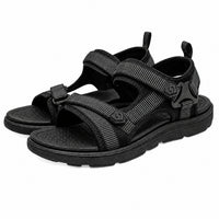 Sandales sport homme avec brides ajustables noires. La chaussure présente une semelle plate très épaisse en caoutchouc noir texturé antidérapant, plusieurs lanières en tissu technique noir avec motifs de lignes, deux fermetures velcro/scratchs réglables en tissu noir gris, des détails de logos gravés en noir sur les scratchs, une bride arrière en tissu avec anse de traction noire, doublure intérieure noire aérée avec perforations, d