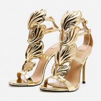Sandales À Talons Hauts Dorées Avec Détails Ailes Sculptées avec finition métallisée dorée, ornements en forme d’ailes, brides fines, bout ouvert et talons aiguilles pour un style luxueux et élégant.